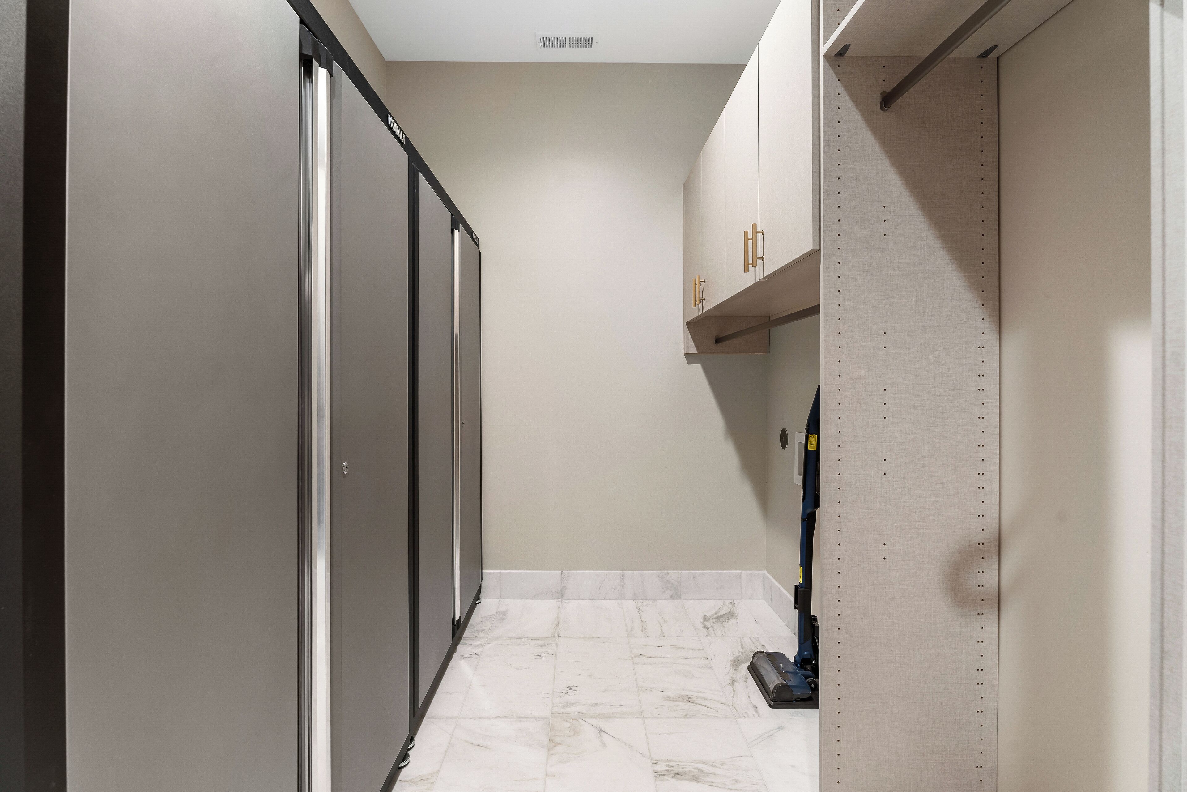 Un couloir étroit, avec une porte coulissante sur la gauche, un aspirateur sur la droite et un sol carrelé de carreaux blancs.