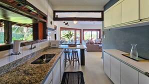 A modern kitchen with granite countertops, a sink, and bar stools. There is a dining area with a table and chairs, and a view of greenery outside. - Casa Maritacas (Sao Sebastiao)