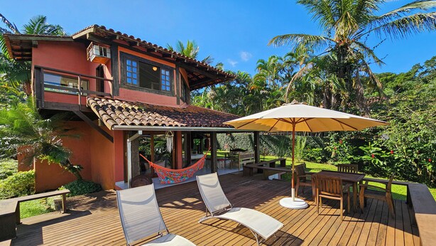 A wooden deck with lounge chairs and a table, a hammock, and a patio area with a view of a house with a red facade and a tiled roof. - Casa Maritacas (Sao Sebastiao)