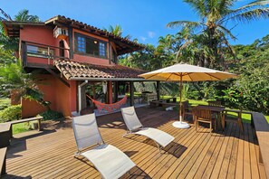 A wooden deck with lounge chairs and a table, a hammock, and a patio area with a view of a house with a red facade and a tiled roof. - Casa Maritacas (Sao Sebastiao)
