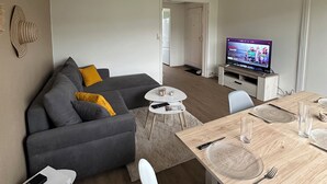 A modern living room with a grey sofa, a small round table, and a dining table with chairs. A television is mounted on the wall, and there is a door leading to another room. - T4 Bright and comfortable - Secure residence (Déville-lès-Rouen)