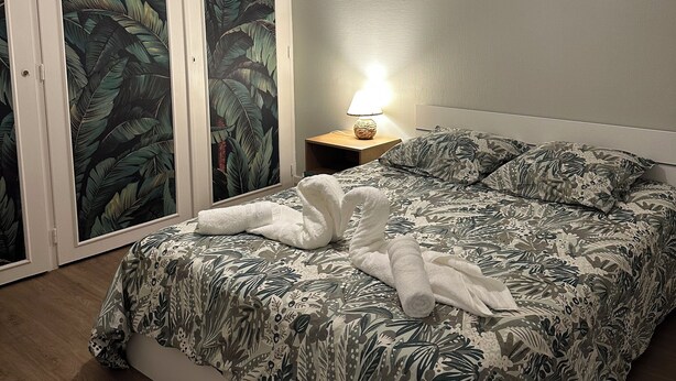 A bedroom with a bed, a nightstand, a lamp, and a door with a leaf-patterned wallpaper. - T4 Bright and comfortable - Secure residence (Déville-lès-Rouen)