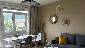 A modern living room with a dining area, featuring a sofa, a dining table with chairs, a coffee table, and a large window with curtains. - T4 Bright and comfortable - Secure residence (Déville-lès-Rouen)