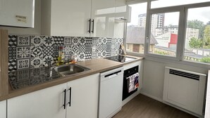 A modern kitchen with white cabinets, a patterned backsplash, a window with a view of buildings, and a ceiling fan. - T4 Bright and comfortable - Secure residence (Déville-lès-Rouen)