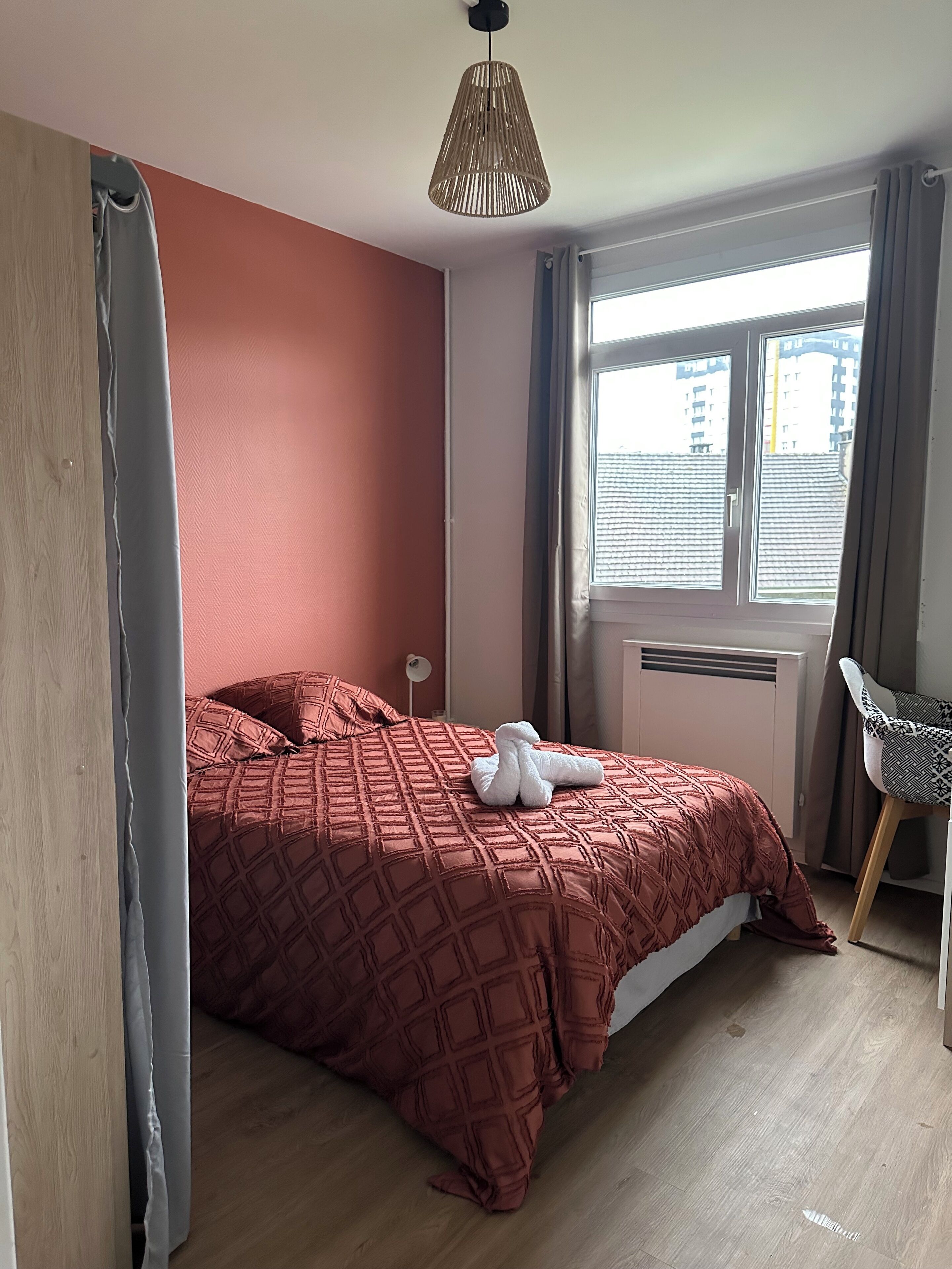 A bedroom with a bed, a window with curtains, a chair, and a hanging light fixture.