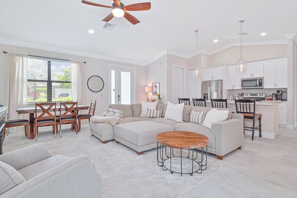 A spacious living room with a sectional sofa, a dining table with chairs, a kitchen area with modern appliances, and a ceiling fan.