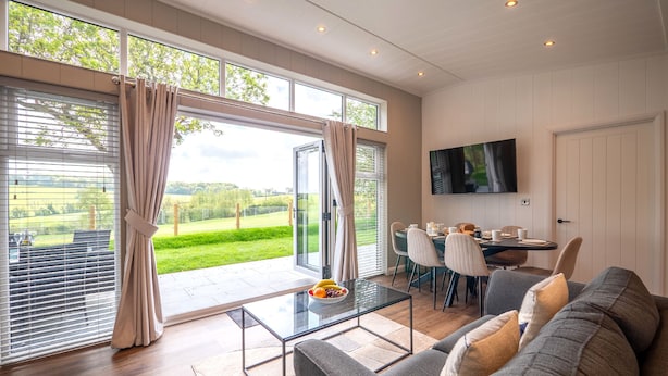 A modern living room with a glass coffee table, grey sofa, and a dining area with a TV mounted on the wall. - A Stylish Retreat Set in a Scenic Country Club (Blackawton)