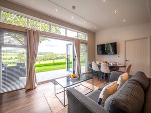 A modern living room with a glass coffee table, grey sofa, and a dining area with a TV mounted on the wall. - A Stylish Retreat Set in a Scenic Country Club (Blackawton)