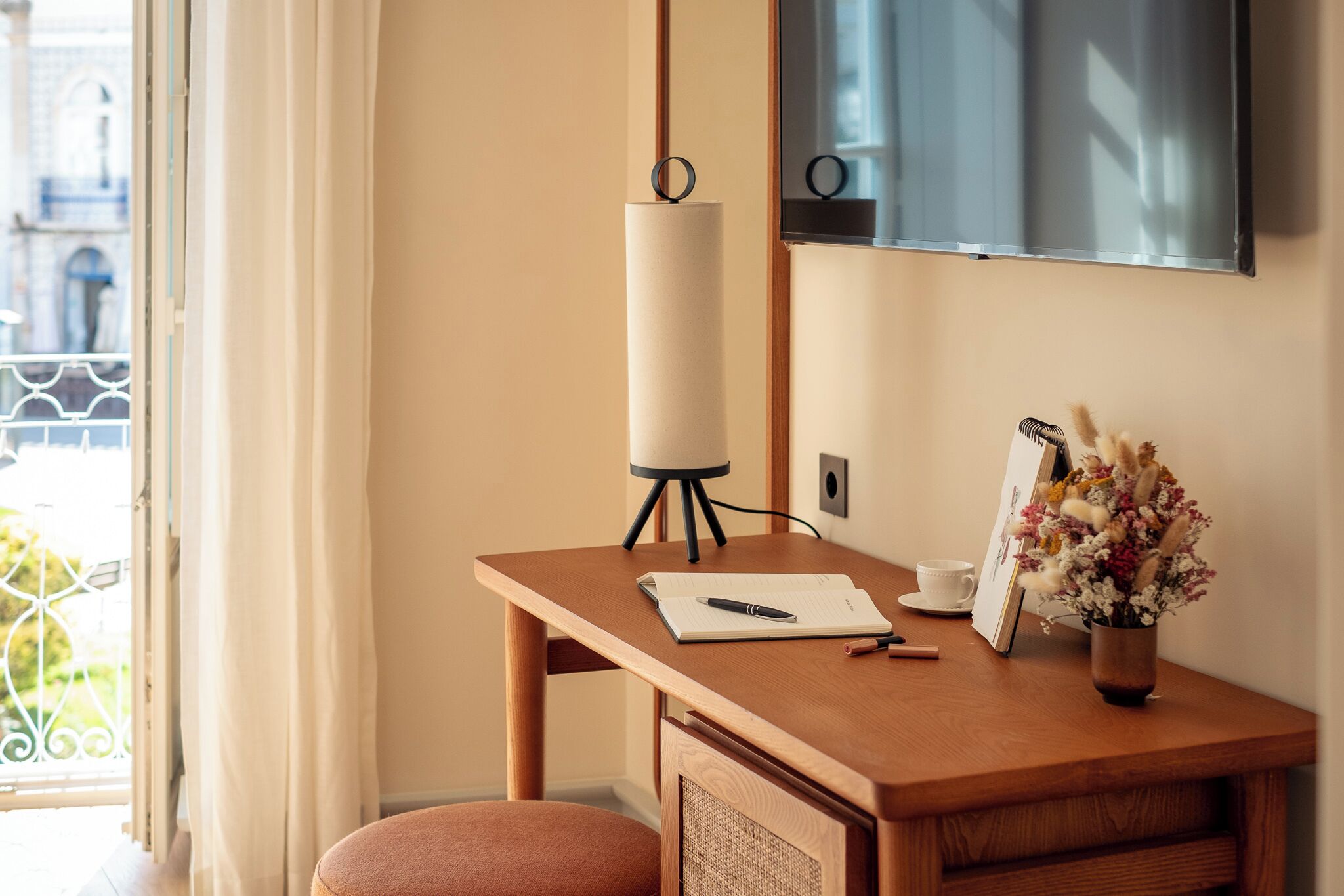 Et hotelværelse med skrivebord i træ, lampe, vase med blomster, billedramme og en kop.