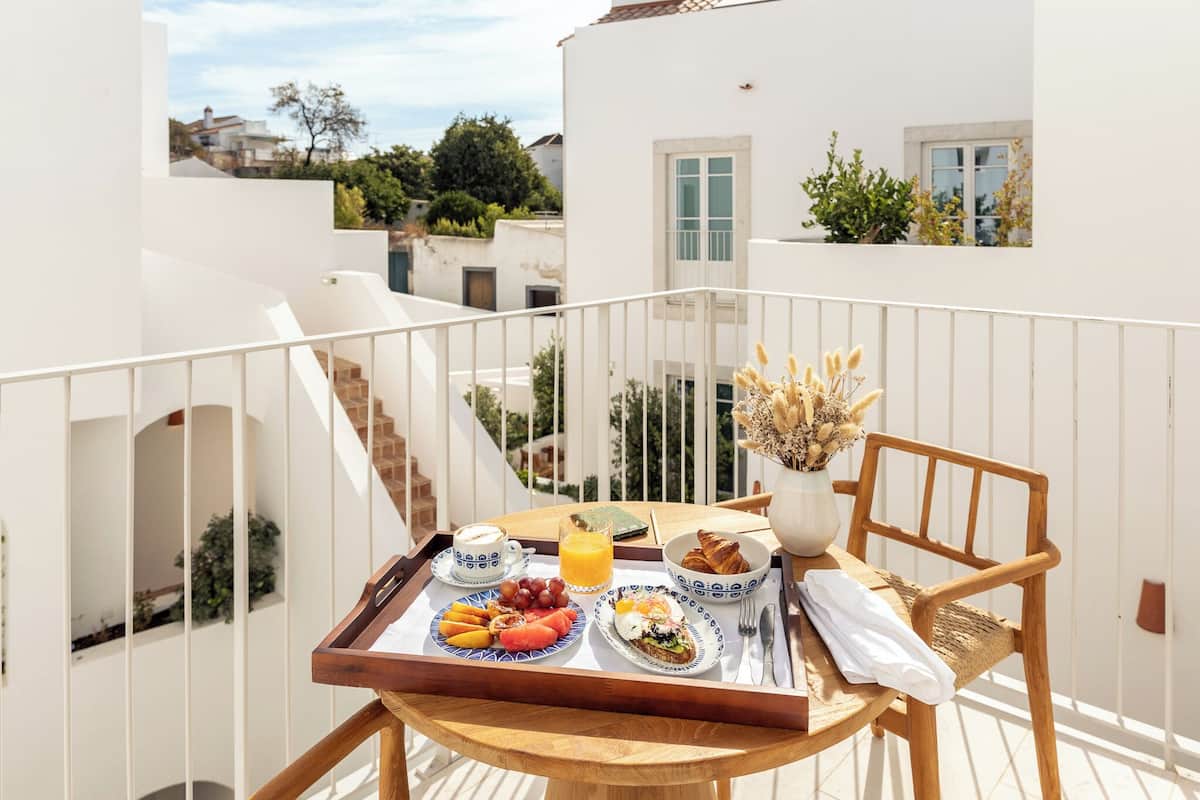 Un balcón con una bandeja de desayuno que contiene croissants, fruta, huevos y jugo, un florero con flores secas y una silla de madera.