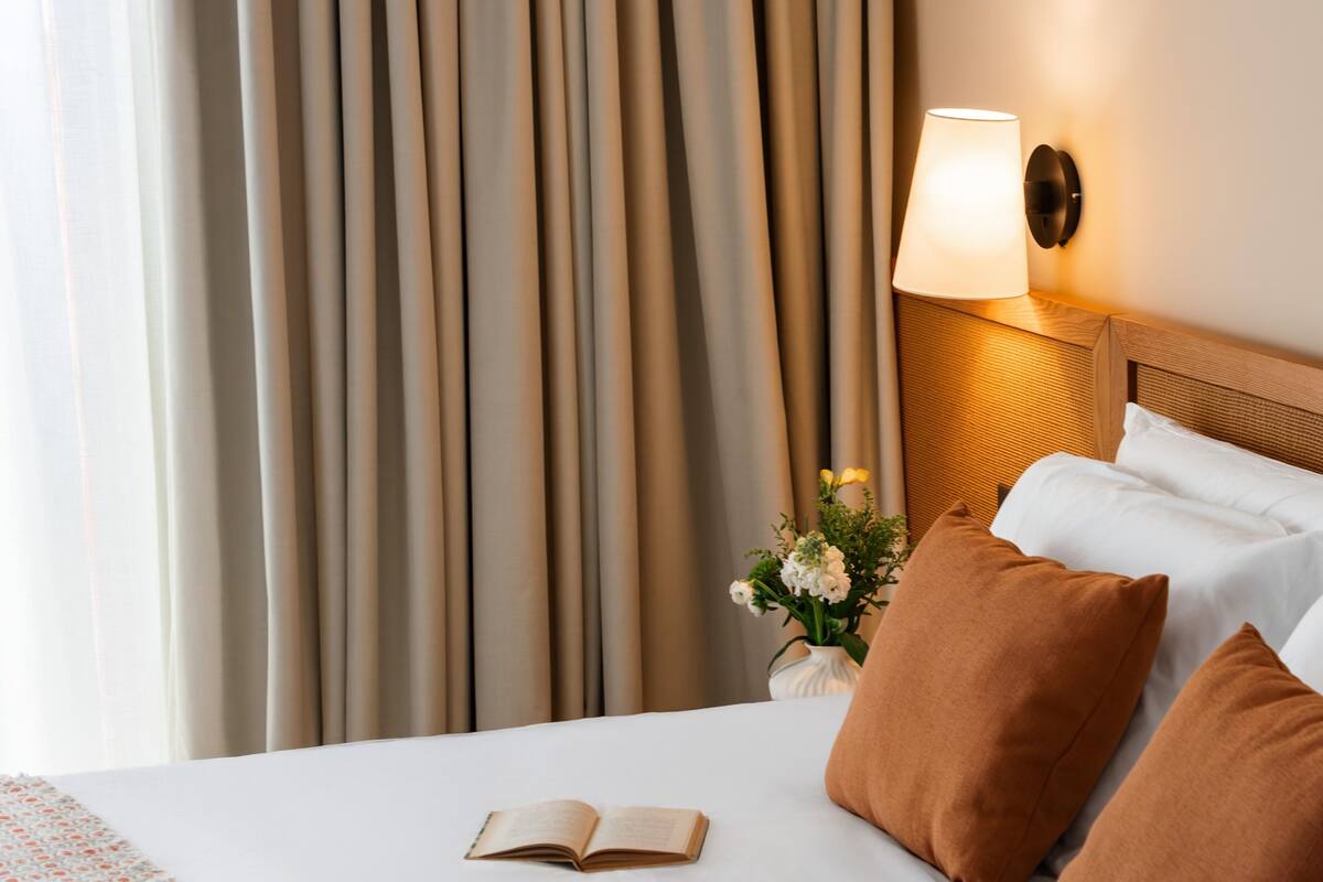 Habitación de hotel con una cama bien hecha, una lámpara de noche, un florero con flores y vistas a cortinas.