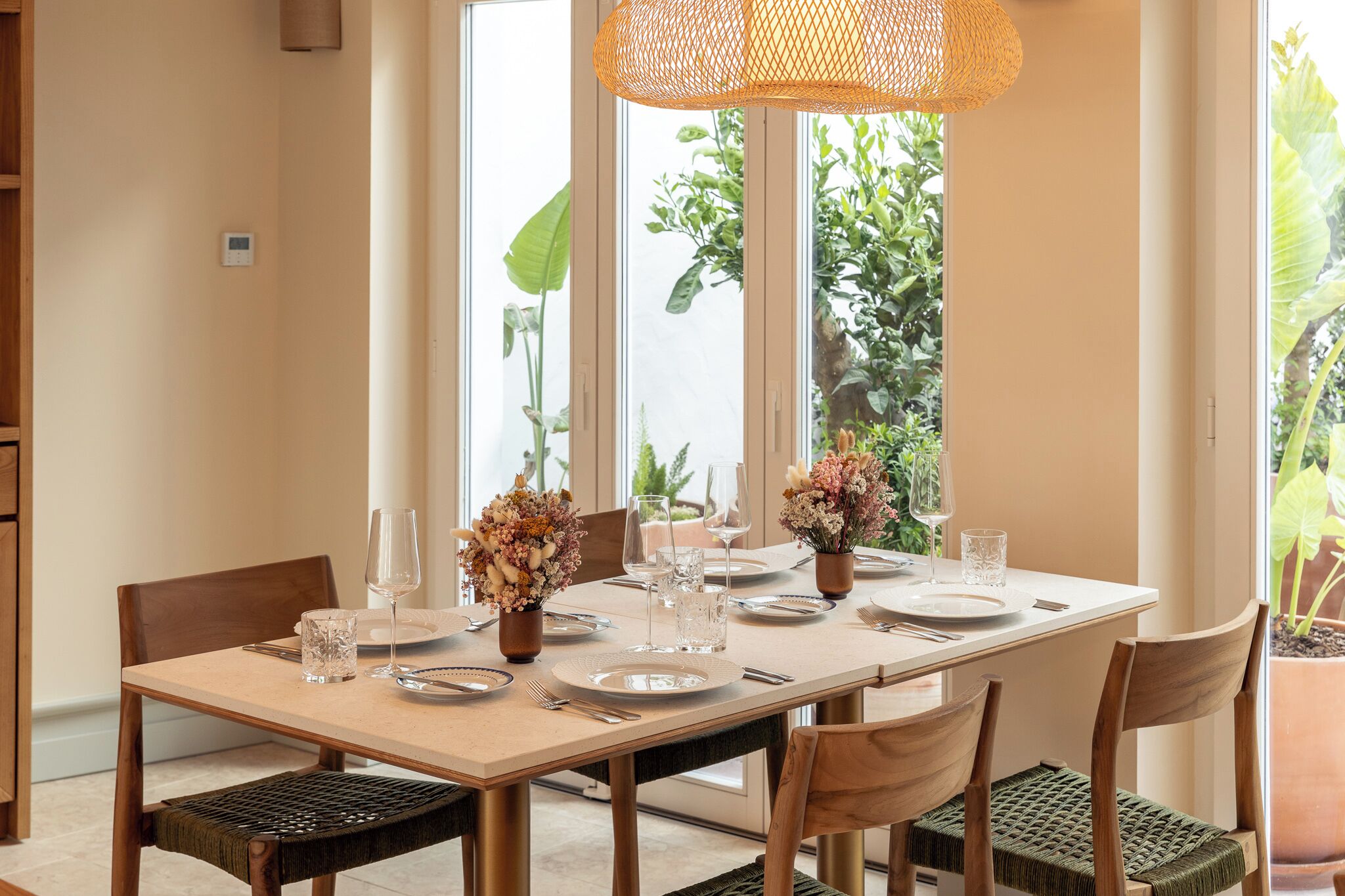 A dining table set with plates, glasses, and cutlery, surrounded by chairs with woven seat cushions.