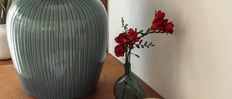 Une table en bois sur laquelle se trouvent une lampe, un carnet, une tasse de café, une assiette de pâtisseries, un bol de raisins et un vase rempli de fleurs rouges.