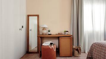 Una camera da letto con un letto, una scrivania in legno, uno specchio, una lampada, un piccolo tavolino con un vaso e un bicchiere, e un cesto di vimini.