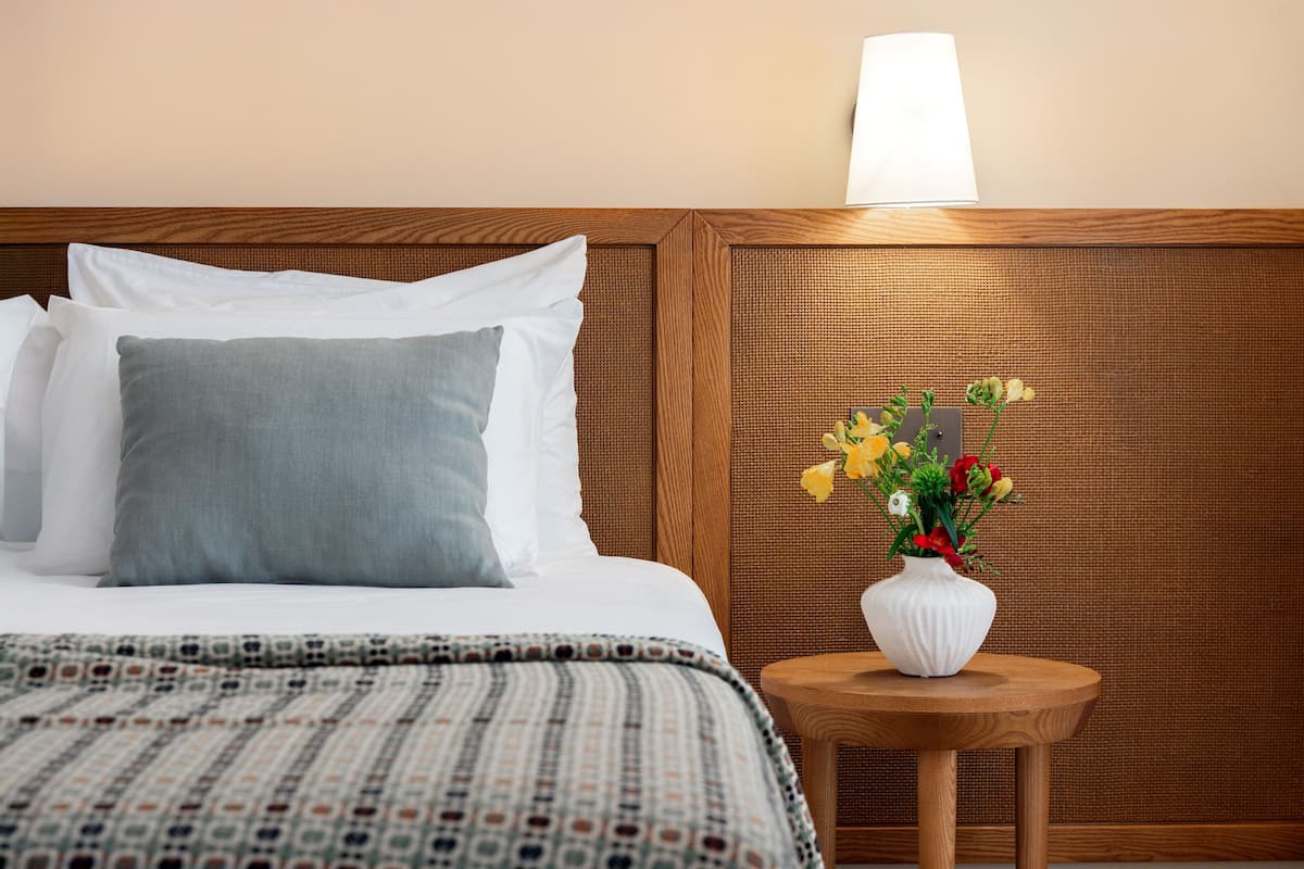 Una cama bien hecha con almohadas blancas y grises, una colcha estampada y una pequeña mesa redonda de madera con un jarrón blanco lleno de flores.