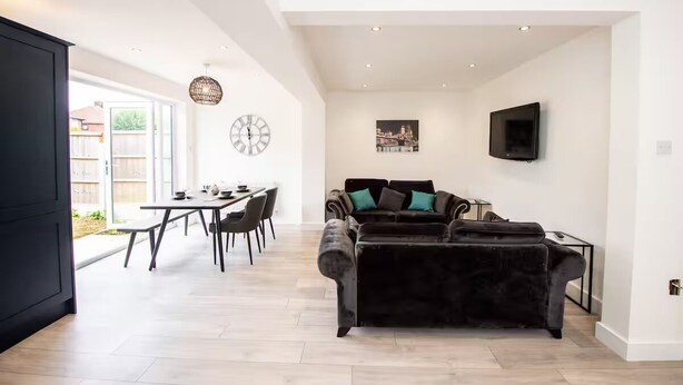 A spacious living area with a dark velvet sofa, a dining table with chairs, a large wall clock, and a flat-screen TV mounted on the wall. - Hillfoot House (Liverpool)
