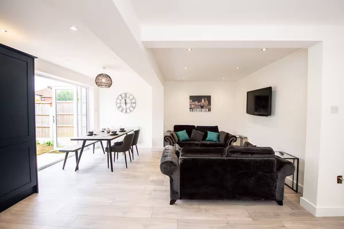 A spacious living area with a dark velvet sofa, a dining table with chairs, a large wall clock, and a flat-screen TV mounted on the wall.