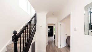 A modern interior with a wooden staircase, a kitchen in the background, and a hallway leading to a door. - Hillfoot House (Liverpool)