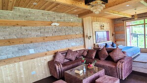 L’intérieur chaleureux d’un chalet avec un plafond en bois, des murs en pierre, un grand canapé en cuir marron, une table basse en verre et un lit recouvert d’une couverture bleue.