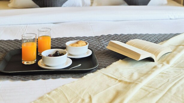 Un lit bien fait, avec des oreillers, une table d’appoint avec un petit-déjeuner comprenant des croissants, un livre et des verres de jus d’orange.