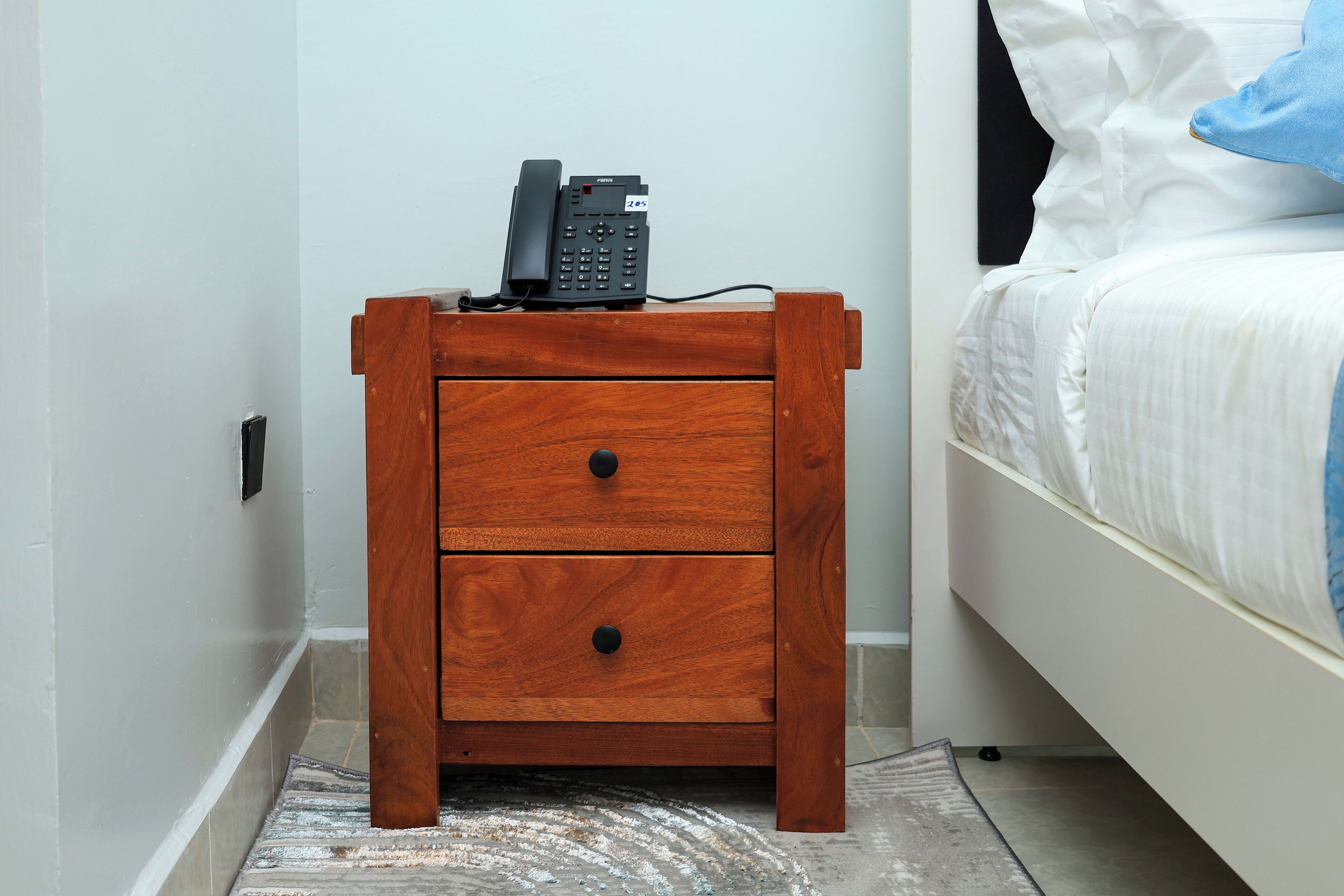 Una mesita de noche de madera con un teléfono y un cajón, junto a una cama con ropa blanca y una almohada azul.