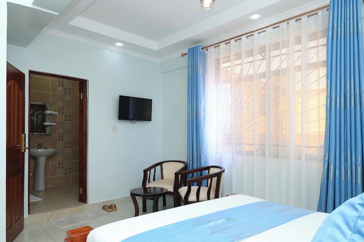Camera d'albergo con un letto, una TV a muro, un tavolino, due sedie, un bagno con un lavandino e una finestra con tende.