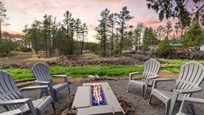 Una hoguera rodeada de cuatro sillas Adirondack, ubicada en un área de grava con vistas a un bosque y casas al fondo.