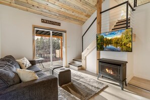 A modern living room with a fireplace, a sofa, a TV mounted above the fireplace, and a staircase leading to an upper level. - Shoreline Village Cabin 3 (Lakeside)