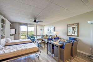 A spacious room with two beds, a dining table, wicker chairs, a ceiling fan, and a large window offering a view of the outdoors. - Steps to Beach & Boardwalk: Condo in Port Aransas! (Port Aransas)