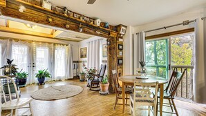 Una habitación amplia con suelo de madera, una mesa redonda para comer y varias sillas. Hay plantas en macetas y un ventanal con vistas a árboles.