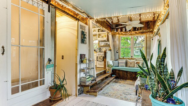 Un ambiente acogedor con un banco de madera, plantas en macetas y una cocina visible a través de una puerta abierta.