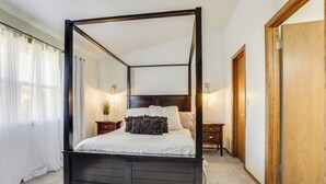 Un lit à baldaquin au revêtement foncé, une couvre-lit blanche et un coussin décoratif. Il y a deux tables de chevet avec des lampes, une porte en bois et un tapis blanc et moelleux sur le sol.