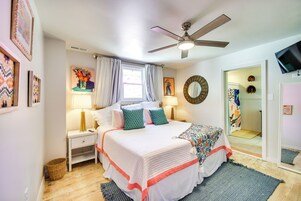 Un dormitorio con cama, mesitas de noche, ventilador de techo, ventana con cortinas y puerta que da a otra habitación.