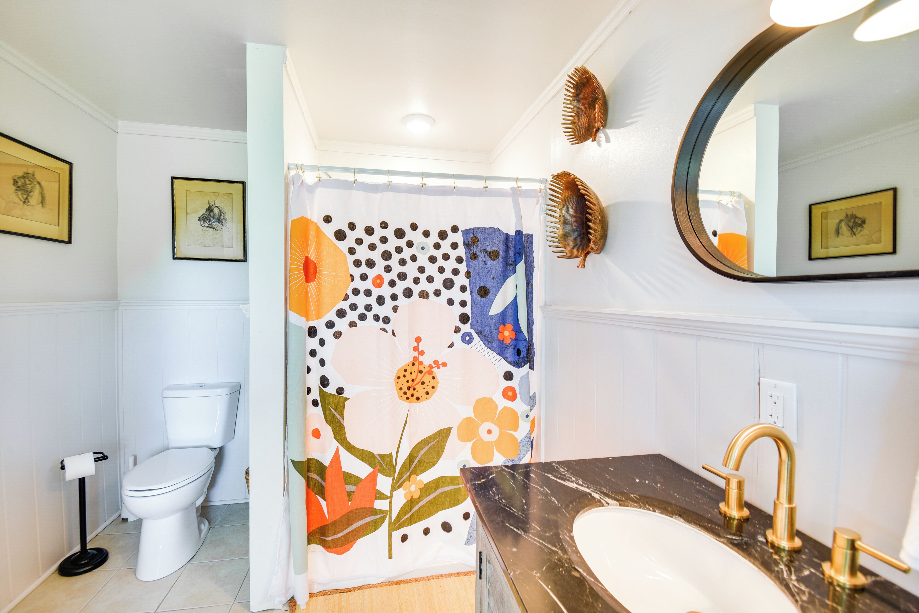 Une salle de bain comprenant des toilettes, un lavabo avec un robinet doré, un rideau de douche à motifs floraux et un miroir au mur.