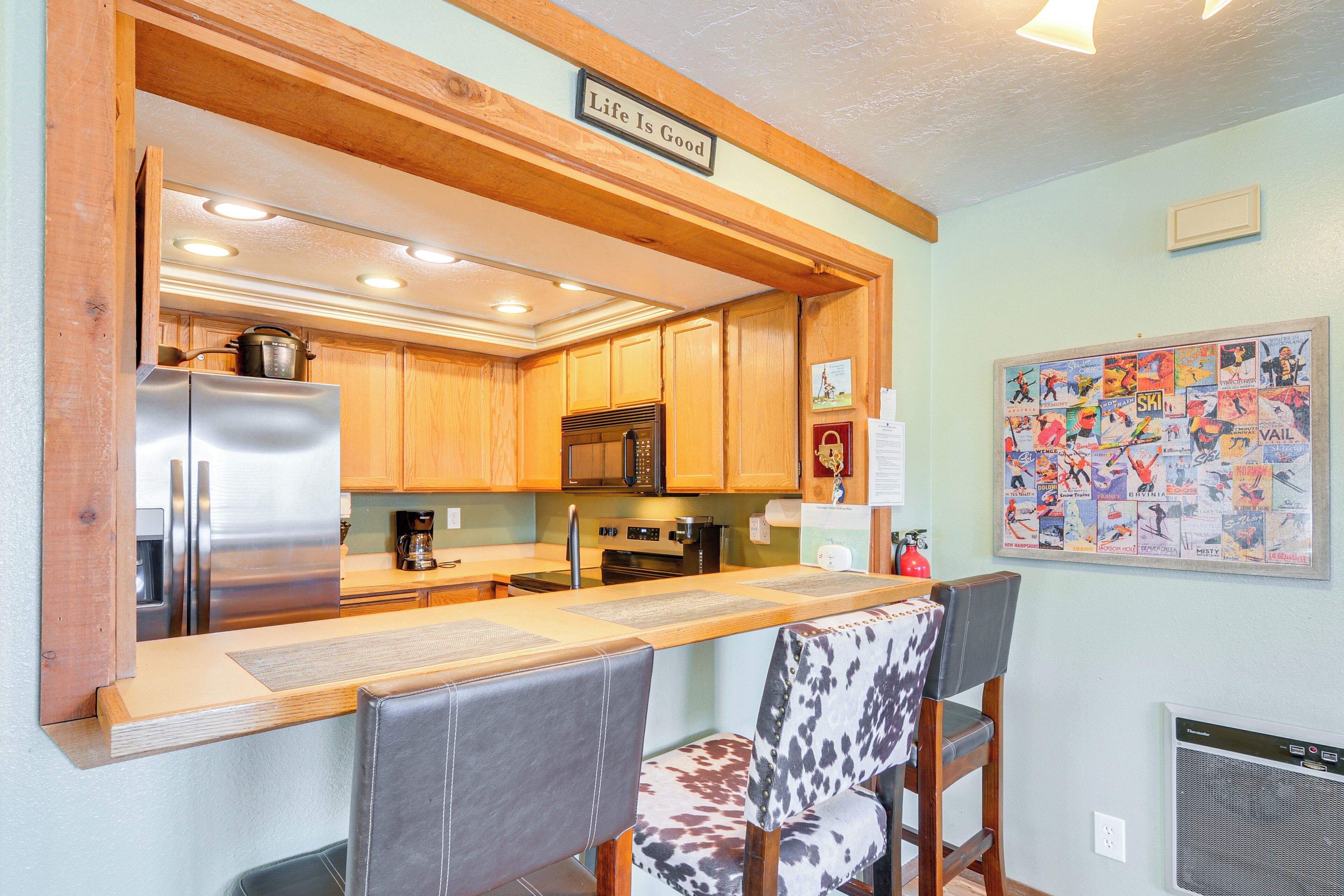 A kitchen with wooden cabinets, a stainless steel refrigerator, a microwave, and a coffee maker. There is a dining area with two chairs and a table. A sign on the wall reads "Life Is Good."
