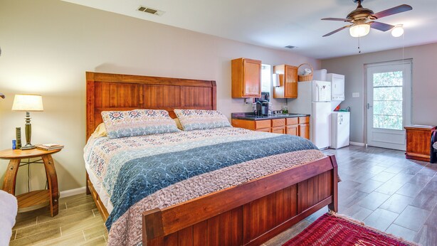 Une chambre avec un lit en bois, une table de chevet, une lampe, un ventilateur de plafond et une vue sur la cuisine.