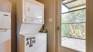 Une buanderie équipée d’un lave-linge et d’un sèche-linge à chargement frontal, d’un panier à linge et d’une porte donnant sur un espace recouvert de gravier et agrémenté d’arbres.
