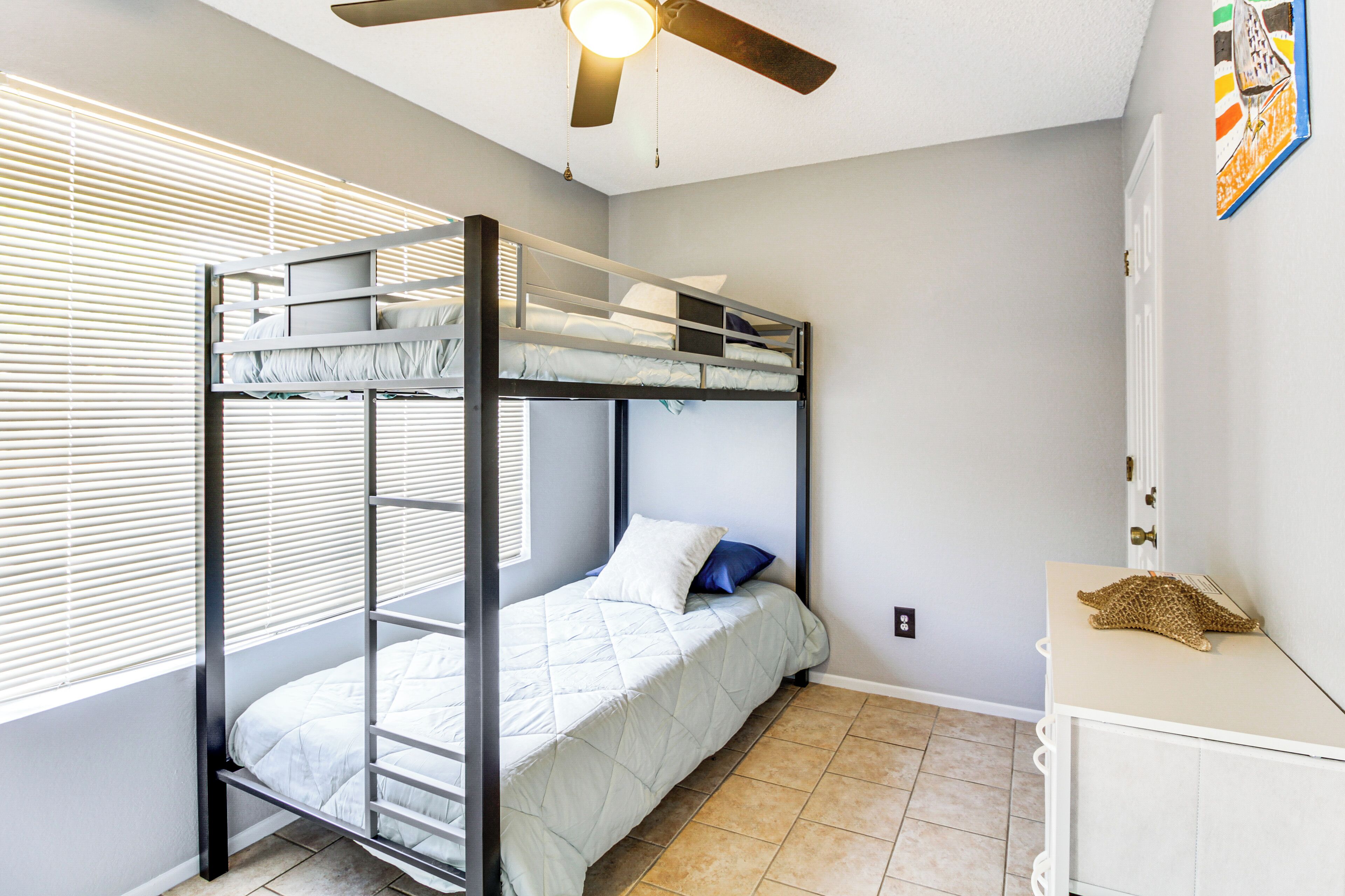 A bunk bed room with a ceiling fan, a window with blinds, and a small cabinet with a decorative item on top.