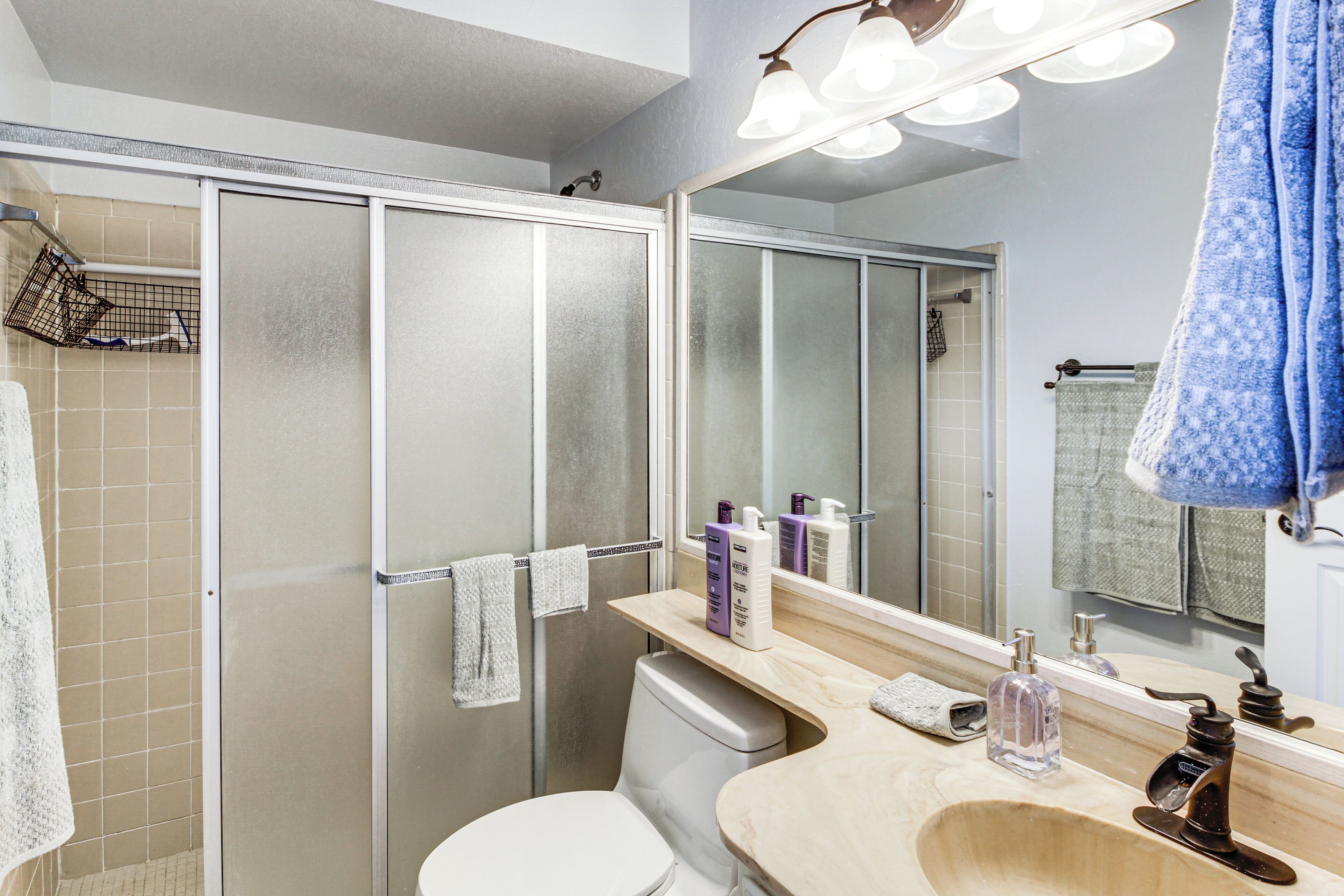 Un baño moderno con cabina de ducha de vidrio, inodoro, lavabo con espejo y diversos artículos de aseo.