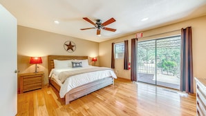 A bedroom with a wooden floor, a bed with white bedding, a ceiling fan, a nightstand with a lamp, and a view through a large window. - Spacious Ruidoso Home w/ Patio & Mountain Views! (Ruidoso)