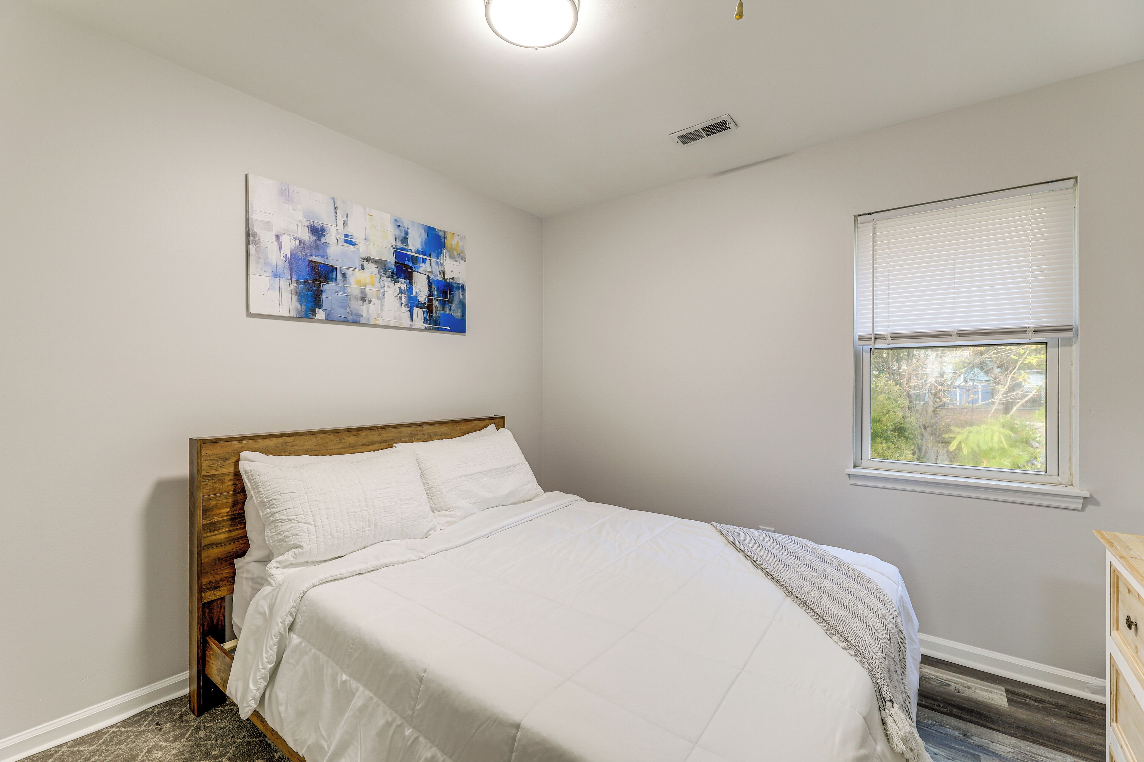 A bedroom with a bed, a window with blinds, a ceiling light, and a wall-mounted abstract painting.