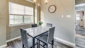 A dining area with a white table, four grey chairs, a chandelier, a clock, and a window with blinds.