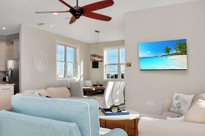 A spacious living room with a ceiling fan, a large TV displaying a beach scene, and a dining area with natural light.