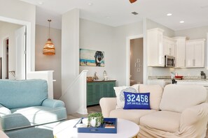A modern living room with a blue sofa, a beige sofa, and a white coffee table. There is a painting on the wall and a kitchen area with white cabinets and a microwave.