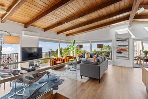Un vaste espace de vie doté d’un plafond en bois, de grandes fenêtres, d’un canapé gris, de fauteuils orange et d’un téléviseur.