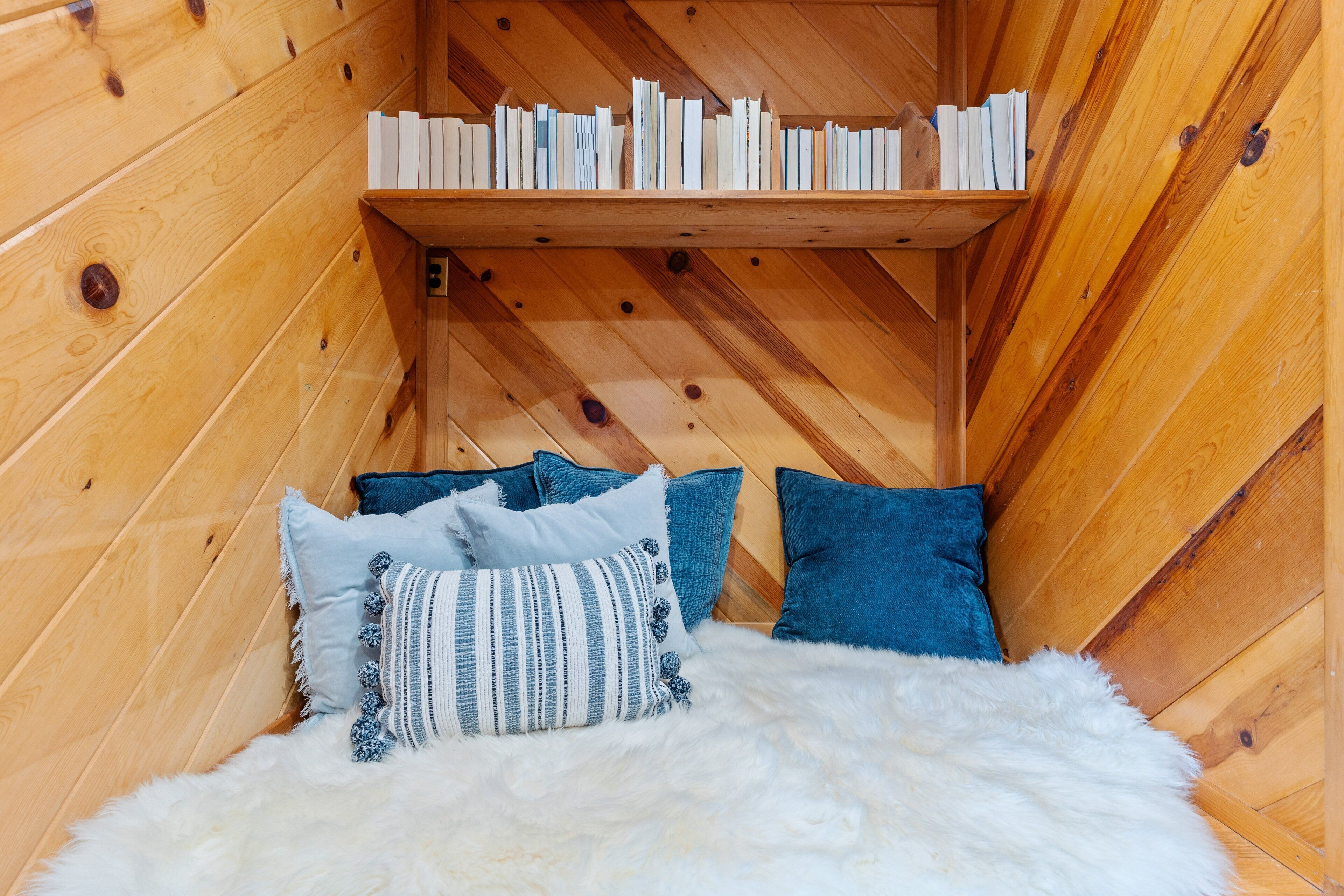 A cozy corner with a wooden shelf holding books, a fluffy white rug, and several pillows.
