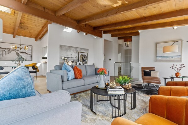A modern living room with a grey sofa, orange armchairs, and a coffee table. There is a painting on the wall and a wooden ceiling. - 180-Degree Ocean Views Deck + Hillside (Half Moon Bay)