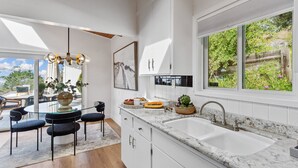 A modern kitchen with a large window, white cabinetry, and a dining area with a glass table and chairs. - 180-Degree Ocean Views Deck + Hillside (Half Moon Bay)