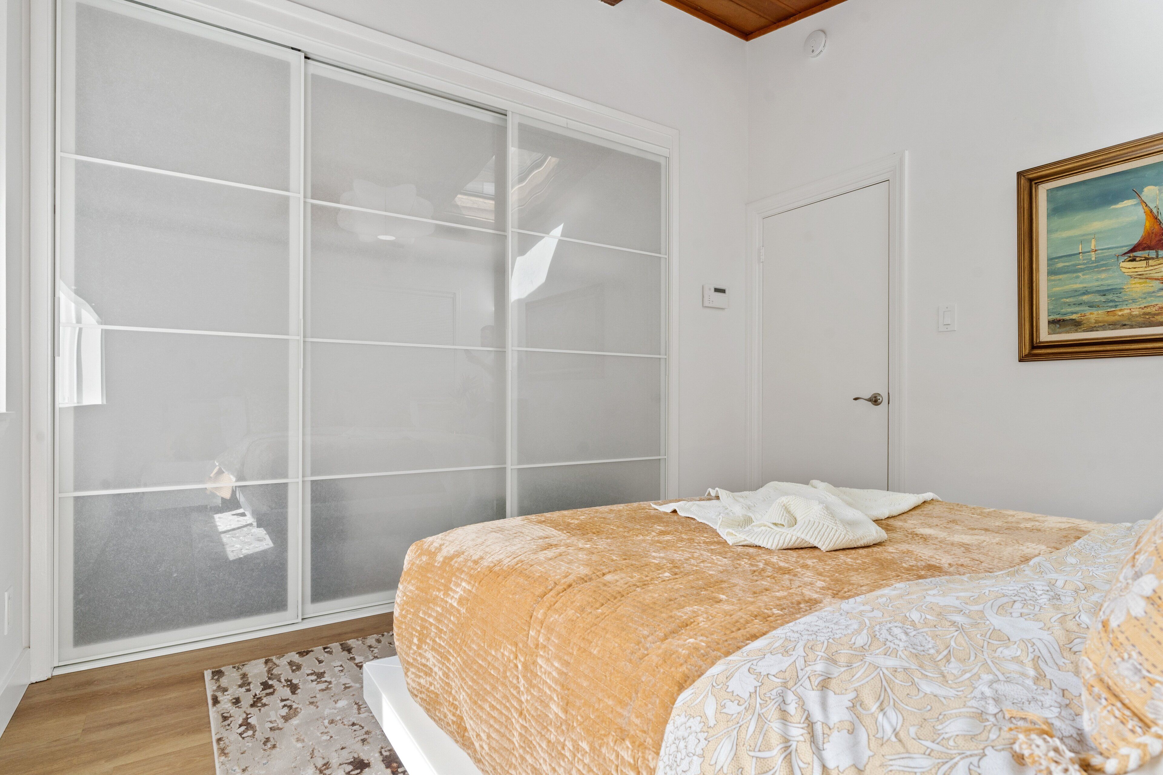 A bedroom with a bed, a large sliding glass door, a painting on the wall, and a wooden floor.