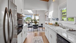 A modern kitchen with white cabinetry, a stainless steel refrigerator, a dining area with a glass table and chairs, and a large window providing a view of greenery outside. - 180-Degree Ocean Views Deck + Hillside (Half Moon Bay)
